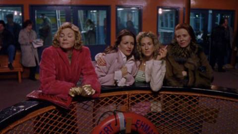 A still of Sex and the City. Carrie, Miranda, Charlotte, and Samantha ride a ferry. They are all bundled up and Carrie is smoking a cigarette. 