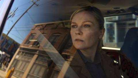 Photojournalist Lee Miller (Kirsten Dunst) sits in a car and pensively looks out of the window at the small town reflected in the glass in Alex Garland’s Civil War
