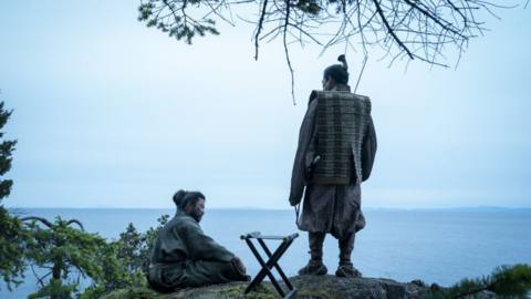 Toranaga (Hiroyuki Sanada) stands next to Tadanobu Asano as Kashigi Yabushige, who is sitting on the ground on a cliffside 