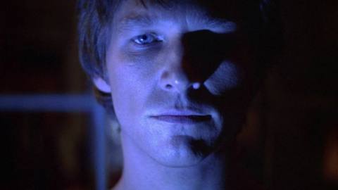 A close-up shot of a man’s stoic face bathed in an unearthly blue light.