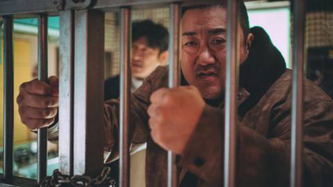 Ma Dong-seok’s massive hands grip the bars of a jail cell as he looks through it in The Roundup: Punishment