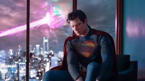 A promotional photo of David Corenswet as Superman, sitting on a mid-century style chair in his uniform, pulling on his boots, as some huge strange energy problem is visible out the window behind him. His costume and boots are textured, but bear the classic Superman coloring, the traditional red S, and a belt with red trunks. 