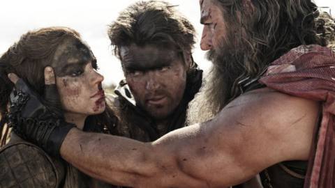 Furiosa (Anya Taylor-Joy), Praetorian Jack (Tom Burke), and Dementus (Chris Hemsworth), three extremely filthy Wasteland-dwellers with greasepaint-smeared faces, huddle close together, with Dementus holding Furiosa’s proud face in his hands, in George Miller’s Furiosa