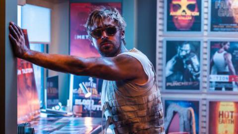 In a scene from The Fall Guy, stunt man Colt Seavers (Ryan Gosling, in sunglasses, a filthy sleeveless vest, and a scowl) leans one-armed against a wall in a room lined with neon-colored movie posters