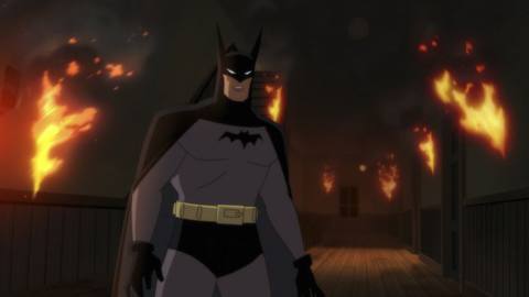 Batman as he appears in the Prime Video series Batman: Caped Crusader, with horn like ears and a small bat symbol. He’s standing in front of a fire