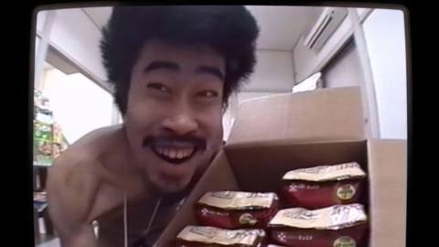 Japanese comedian Nasubi, naked and with a huge pouf of wild hair, grins as he holds up a box full of the bags of dog food he’s about to eat on the game show Susunu! Denpa Shōnen, as seen in the documentary The Contestant
