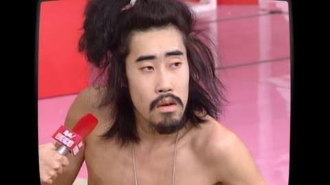 Tomoaki “Nasubi” Hamatsu, a Japanese man with a long face, a moustache, and long hair in a top knot, has a blank expression on his face as he looks away from a TV interviewer’s red microphone, in archival footage from the Japanese game show Susunu! Denpa Shōnen, as seen in the documentary The Contestant
