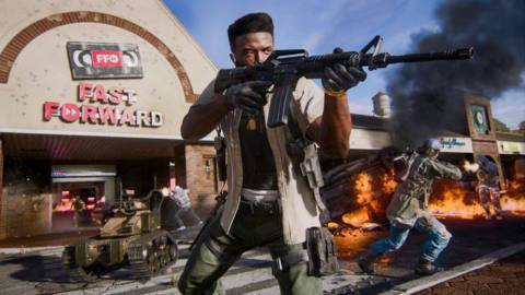 A soldier in civilian clothing stands guard outside of a video rental store named Fast Forward while a fire burns behind him in a screenshot from the Call of Duty: Black Ops 6 multiplayer mode