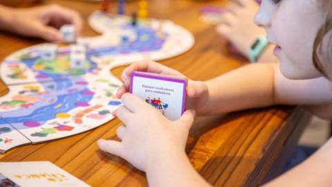 A child sits beside a winding path, laid out as a set of cardboard tiles on the table. She’s holding a small stack of cards. In the background another hand holds a custom die above the bard.