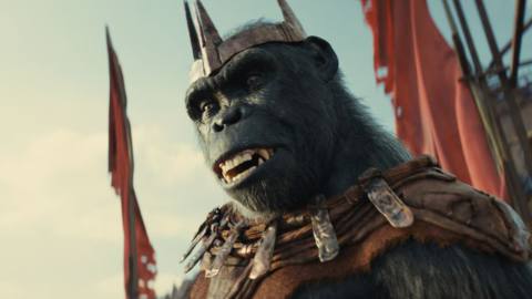 The king Proximus, a black ape in a crown and a heavy necklace, stands in front of banners in the film Kingdom of the Planet of the Apes