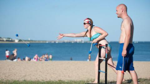 At Carson Beach, in Boston, two beachgoers toss tethered golf balls in a game of Ladderball. The sky is clear, and they’re both wearing swimsuits.