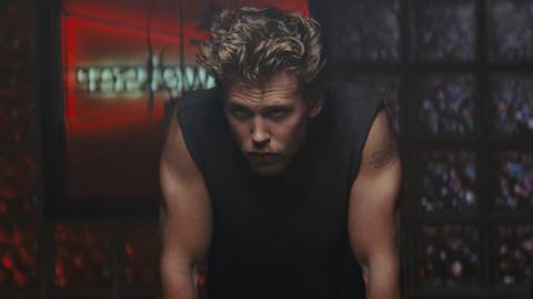 Austin Butler, with mussed-up hair, wearing a black sleeveless top, leans forward in a moody way in The Bikeriders