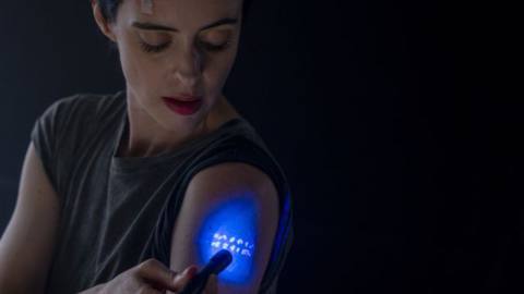 Lucy (Krysten Ritter) holds a blacklight up to her arm to better see some numbers on her skin