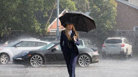 Blake Lively, wearing a dark suit and hat, walks in the rain with an umbrella in A Simple Favor