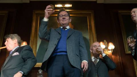 Dr. Leland Townsend (Michael Emerson), flanked by two lawyer looking types with expensive suits, cigars and whiskey, toasts the camera from above in a scene from season 4 of Evil.