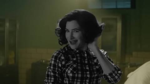 Kathryn Hahn as Agatha/Agnes in Agatha All Along. She smiles and preens her hair, standing in a green tiled room. She, however, appears entirely in black and white, like an old sitcom.