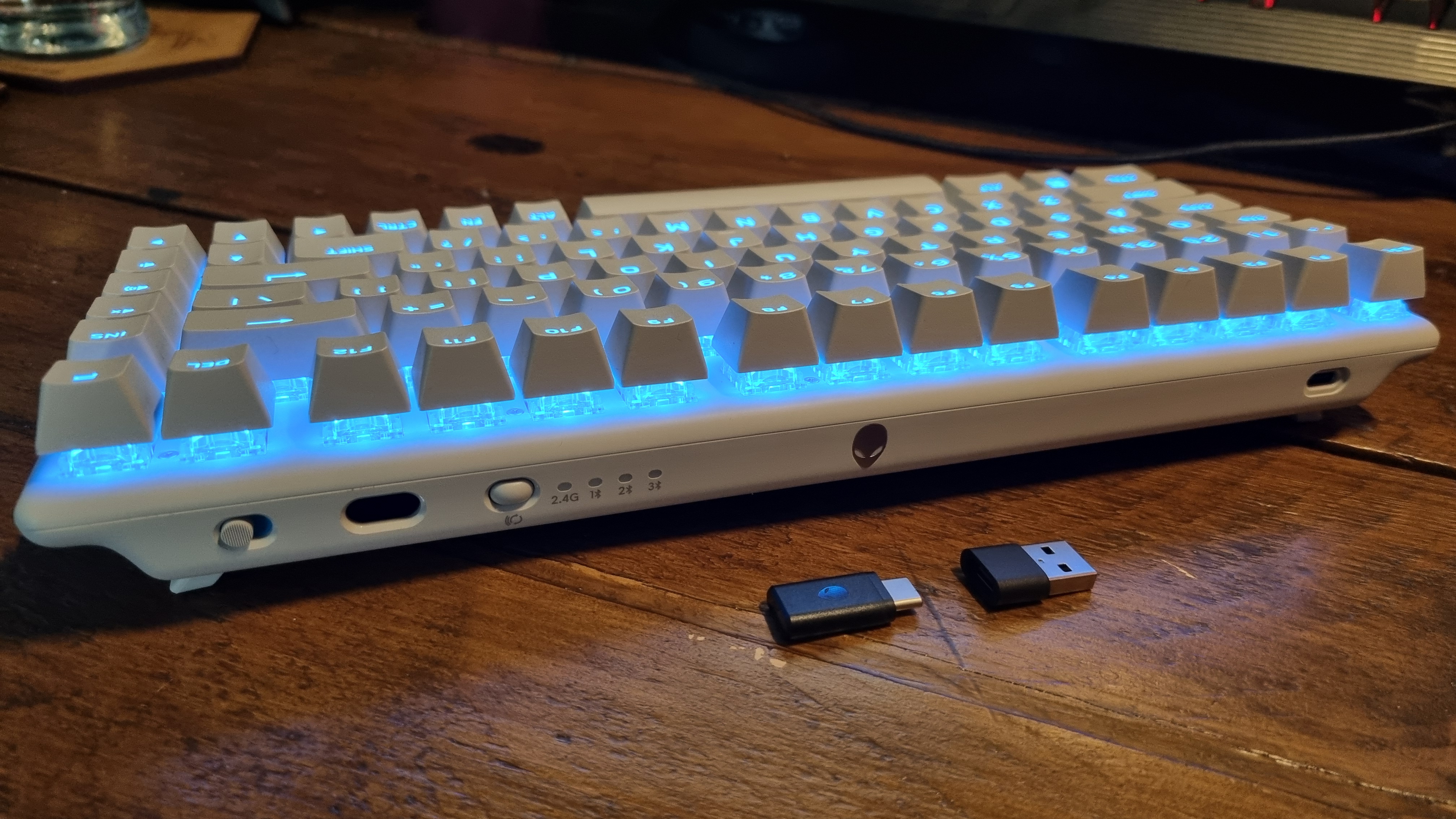 The rear of the Alienware Pro Wireless Gaming Keyboard, lit up in blue, showing the USB dongle slot and associated dongle and adaptor