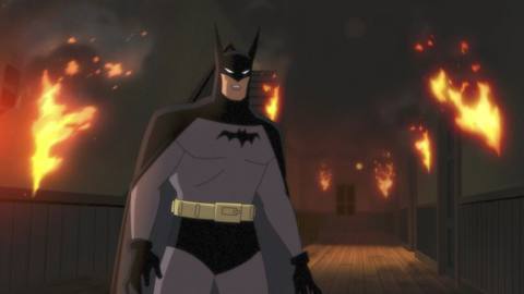 Batman as he appears in the Prime Video series Batman: Caped Crusader, with horn like ears and a small bat symbol. He’s standing in front of a fire