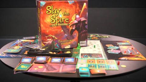 A table covered in cards and player mats from the board game Slay the Spire, with the box for the game sitting up right in the middle.