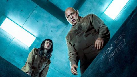 A dark-haired woman and a heavyset balding man in dull brown prison jumpsuits stare down the concrete shaft linking their prison cell to a cell below in a detail from the poster for Netflix’s sci-fi/horror thriller The Platform 2