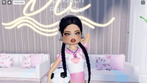 An image of a character in Dress to Impress in Roblox. She weras long pig tail braids, a black and white gingham mini skirt, and a crop top with a pink heart on it. She’s holding a soccer ball and posing for the camera.