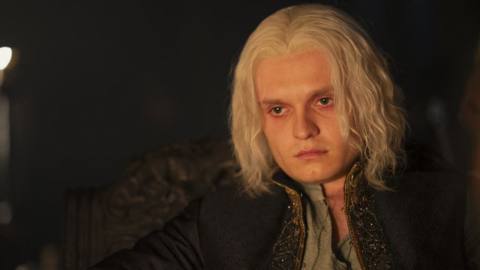 Tom Glynn-Carney as Aegon Targaryen in House of the Dragon, sitting at a table lit by a fire looking upset