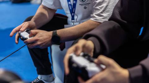 A close-up of two people holding PlayStation 5 controllers at an event.