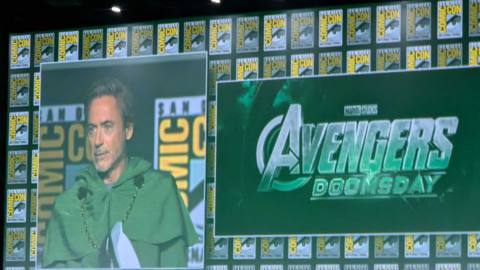 Robert Downey Jr. on a screen wearing green opposite a logo for Avengers: Doomsday, the new MCU movie announced at SDCC 2024