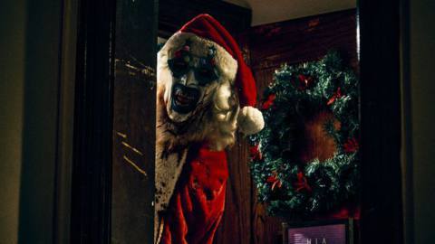 Art the Clown (David Howard Thornton), the signature baddie of the Terrifier season, wears a Santa suit and a dark-painted toothy grin as he looms through a doorway in front of a Christmas wreath in Terrifier 3