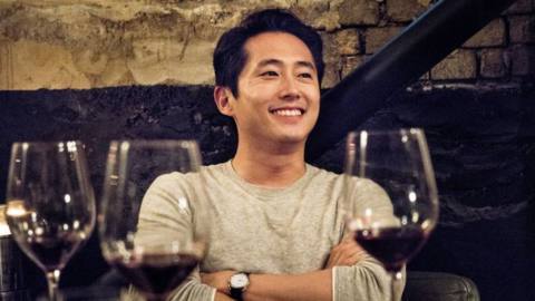 Steven Yeun wearing a white sweater and sitting at a table with wine glasses around him and a big smile on his face in Burning