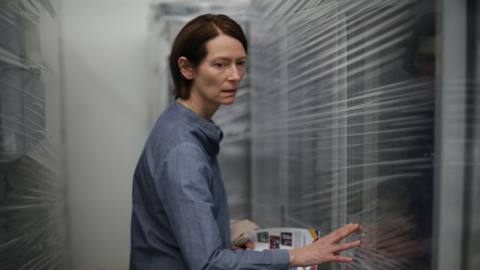 Tilda Swinton looking confused against a blurred background of white slats in Memoria