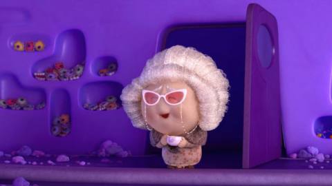 Nostalgia, a character from Pixar’s animated movie Inside Out 2, stands in a doorway. She looks like a little old lady with white hair, and pink cats-eye glasses attached to her face with a chain.