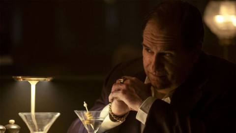 A man with a visible facial scar in a purple suit folding his hands in front of table with cocktail glasses in The Penguin.