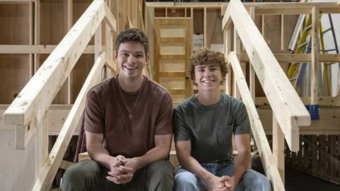 Walker Scolbell and Daniel Diemer sit on a wooden staircase. They are two sandy-haired white boys.