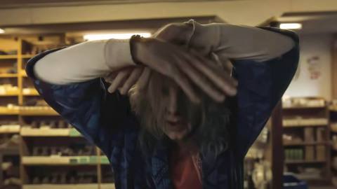 Satanic serial killer Longlegs, a chalk-skinned man with distended, puffy features and long, lank white hair, stands at a hardware store counter, partially covering his face with his hands, in Oz Perkins’ Longlegs