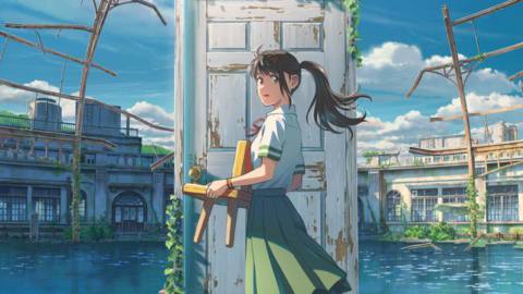 Suzume, a teenage girl in a school uniform, holds a chair as she stands in front of a dilapidated doorway in the middle of a shallow body of water, which itself is in the middle of some ruins
