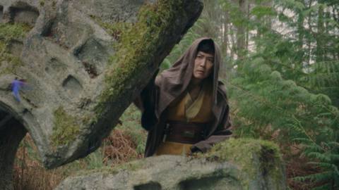 The Jedi Sol stands with his hood up, leaning behind a patterned arch in the woods in a scene from The Acolyte.