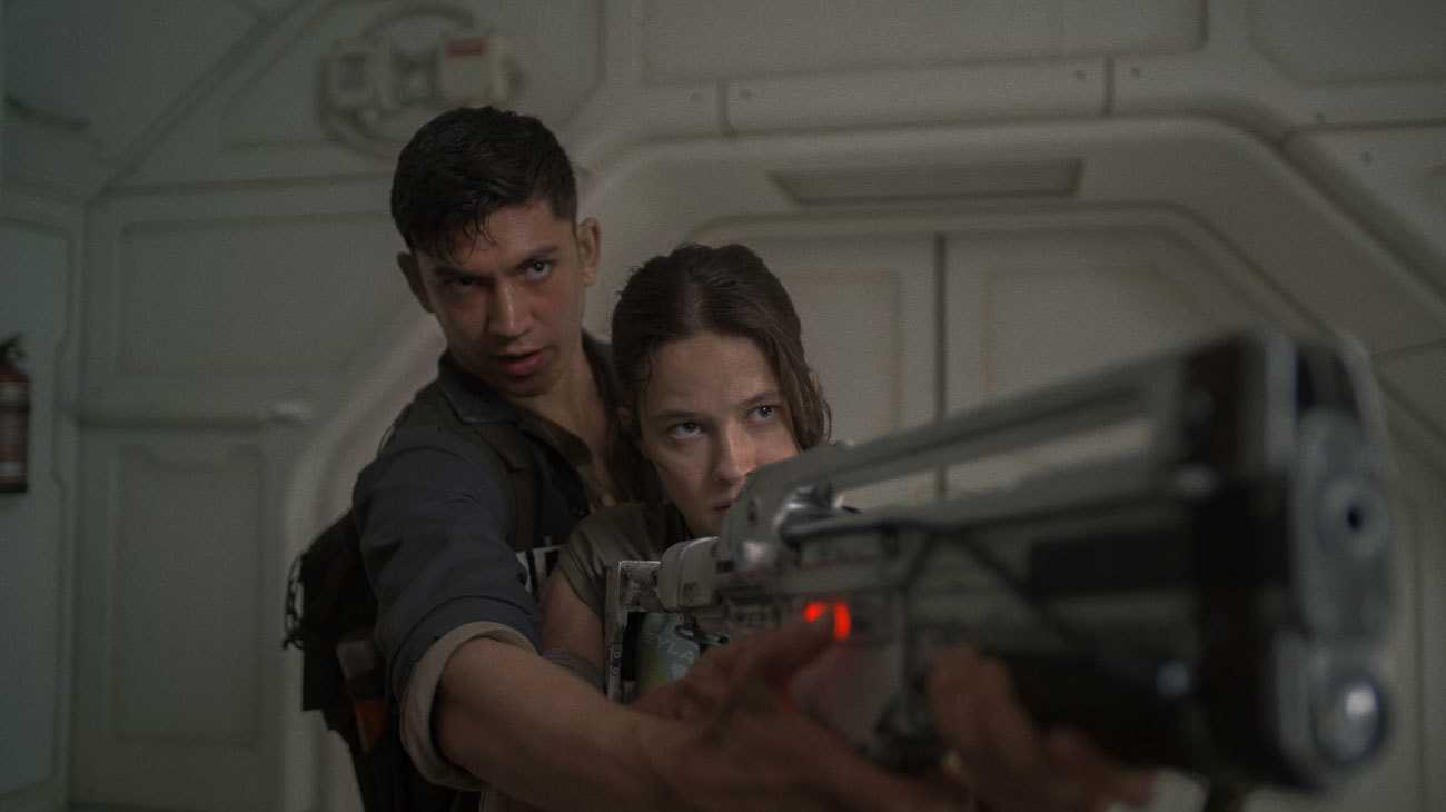 A man standing over the shoulder of a woman as she aims down the sights of a futuristic rifle in Alien: Romulus.A man standing over the shoulder of a woman as she aims down the sights of a futuristic rifle in Alien: Romulus.