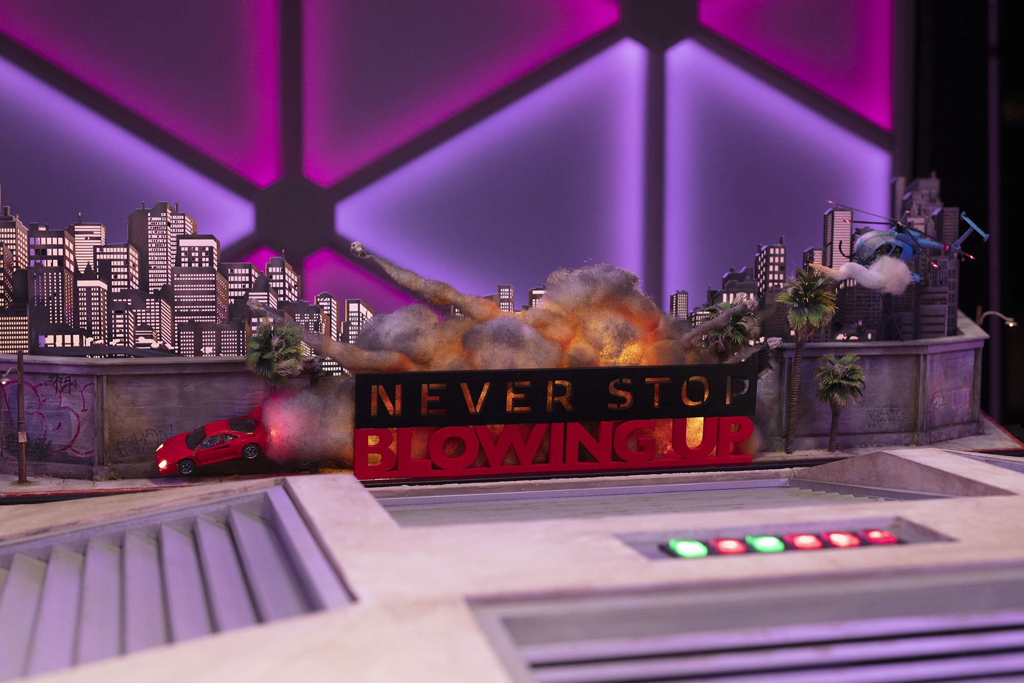 Set design for Dropout’s Never Stop Blowing Up, with a sports car, a city skyline, and explosions