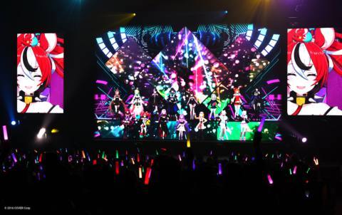 A photo of an in-person hololive concert. There are nine Vtubers on stage and they are performing as holograms. People hold light sticks in the crowd.