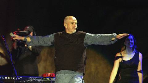 Peter Gabriel wears a bulky tunic on stage, arms stretched out with microphone in hand.