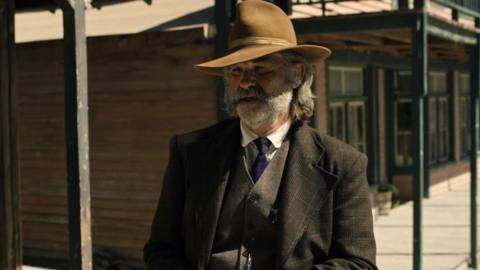 Kurt Russell wearing a cowboy hat and leaning against a wooden post outside a saloon in Bone Tomahawk 