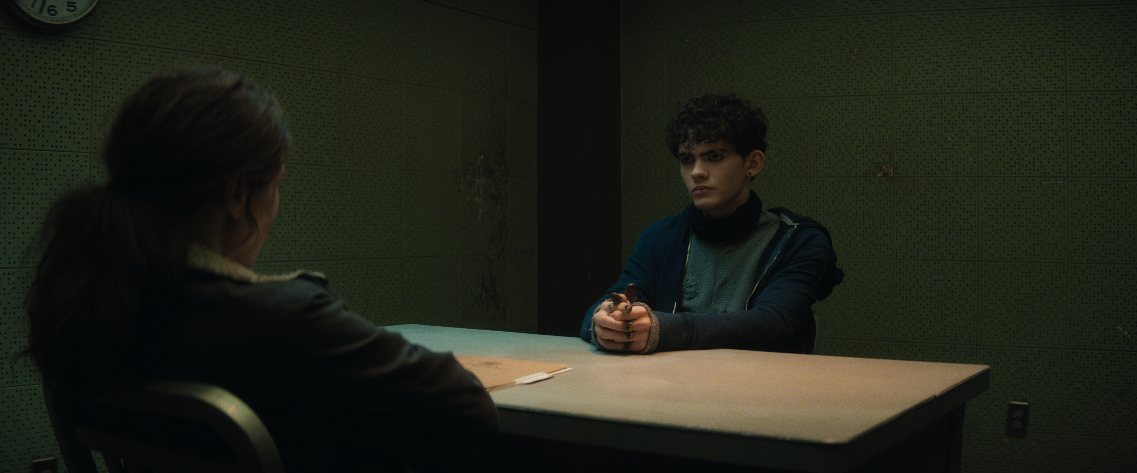 (L-R): Agatha Harkness (Kathryn Hahn) and “Teen” (Joe Locke) in Agatha All Along. Teen is seated at the table in a dark police interrogation room, and Agatha is sitting across from him.