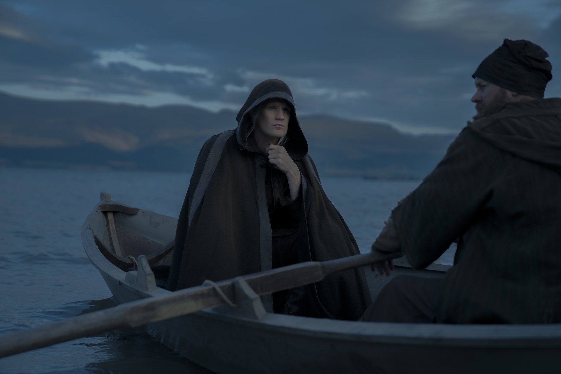 Matt Smith as Daemon Targaryen, wearing a large cloak with a hood and riding in a small boat in House of the Dragon