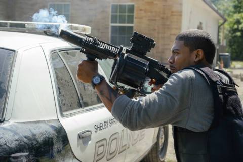 Terry Richmond (Aaron Pierre), a tall Black man with close-cropped hair, wearing a bulletproof vest, crouches behind a police car and fires an immense gun in Netflix’s action movie Rebel Ridge