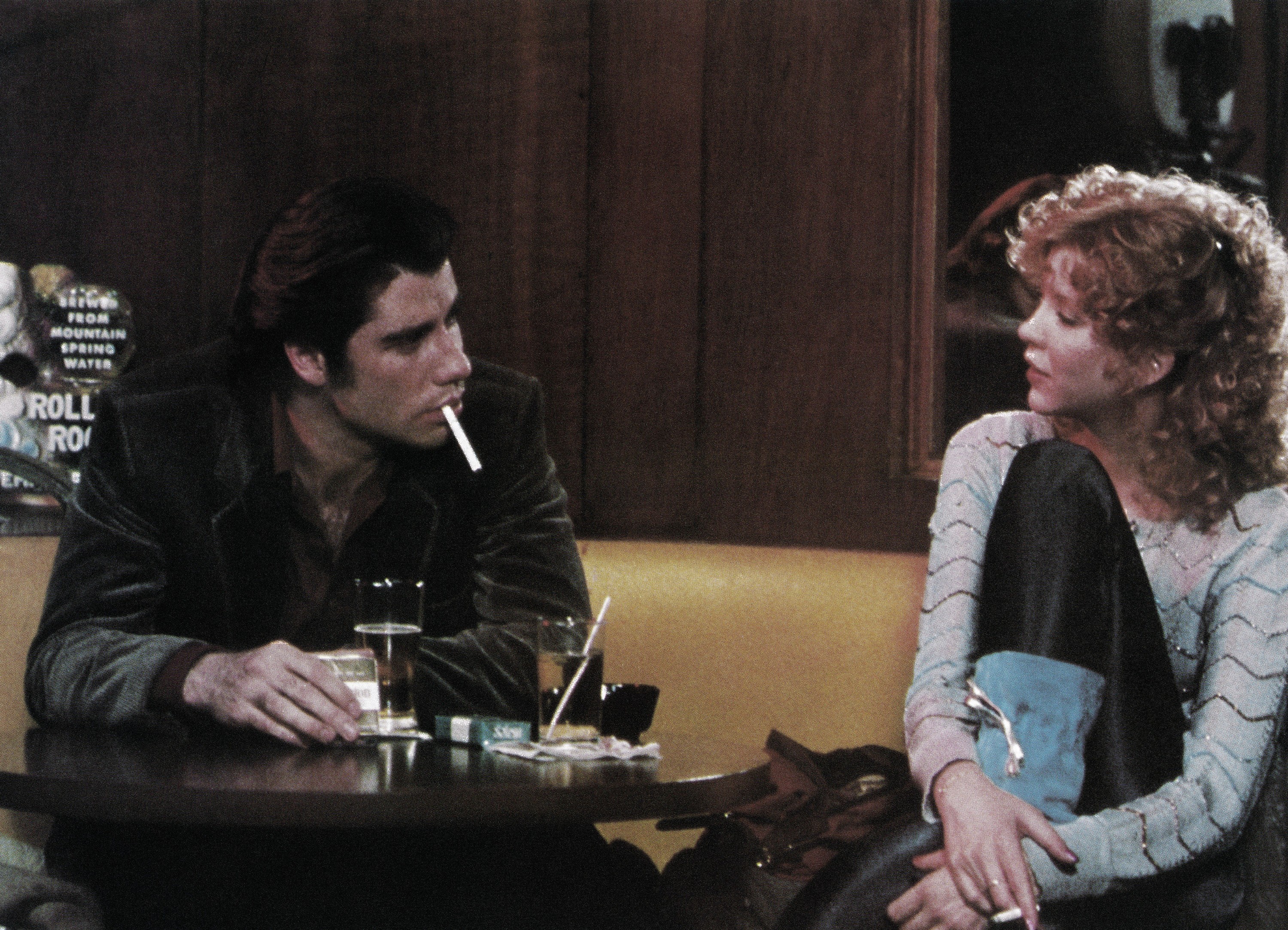 John Travolta and Nancy Allen sit together at a table while Travolta smokes a cigarette in the movie Blow Out