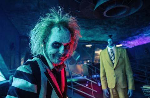 The wild-haired, stripe-suited ghoul Betelgeuse (Michael Keaton) smirks over his shoulder as his shrunken-headed, huge-bodied employee Bob lurks in the background in Beetlejuice Beetlejuice.