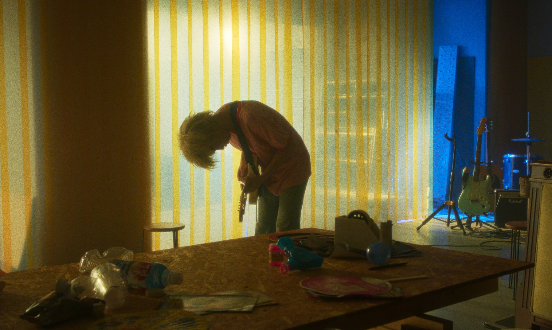 A young man with short bleached hair holding a guitar in an apartment building in Plastic.