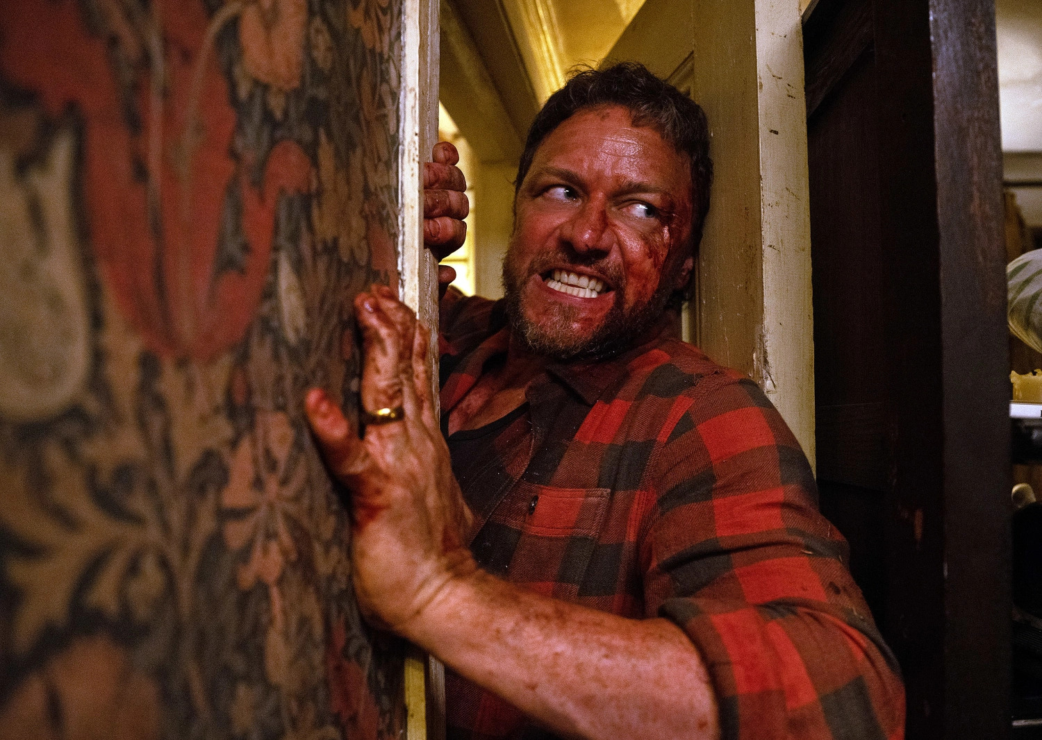 James McAvoy in a red checkered shirt covered with blood and gritting his teeth while pushing his way through a blocked door in Speak No Evil.