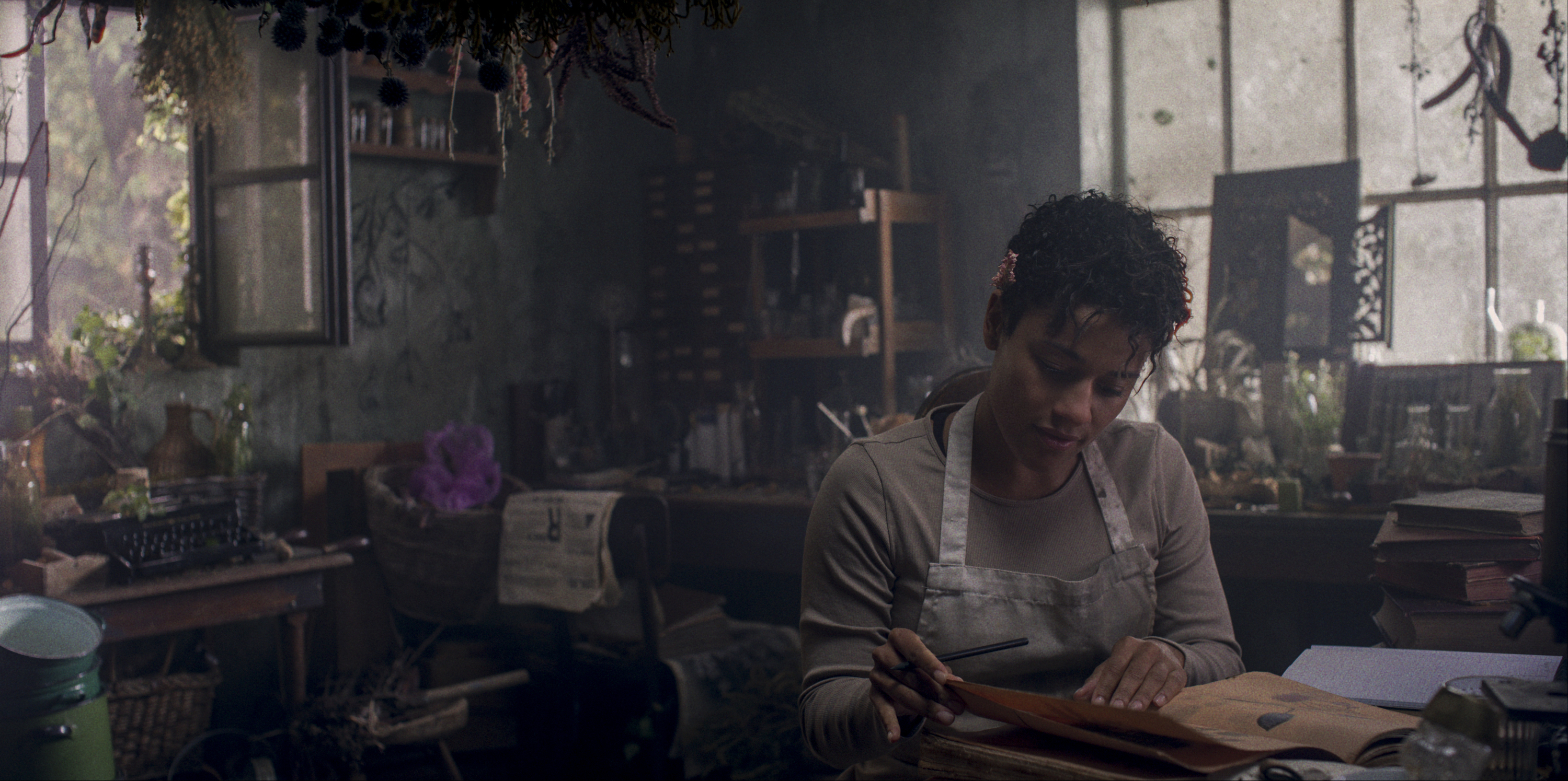 Chef (Ariana DeBose) peruses sheets of parchment and paper in a dark, overgrown, cluttered manor-house kitchen in House of Spoils